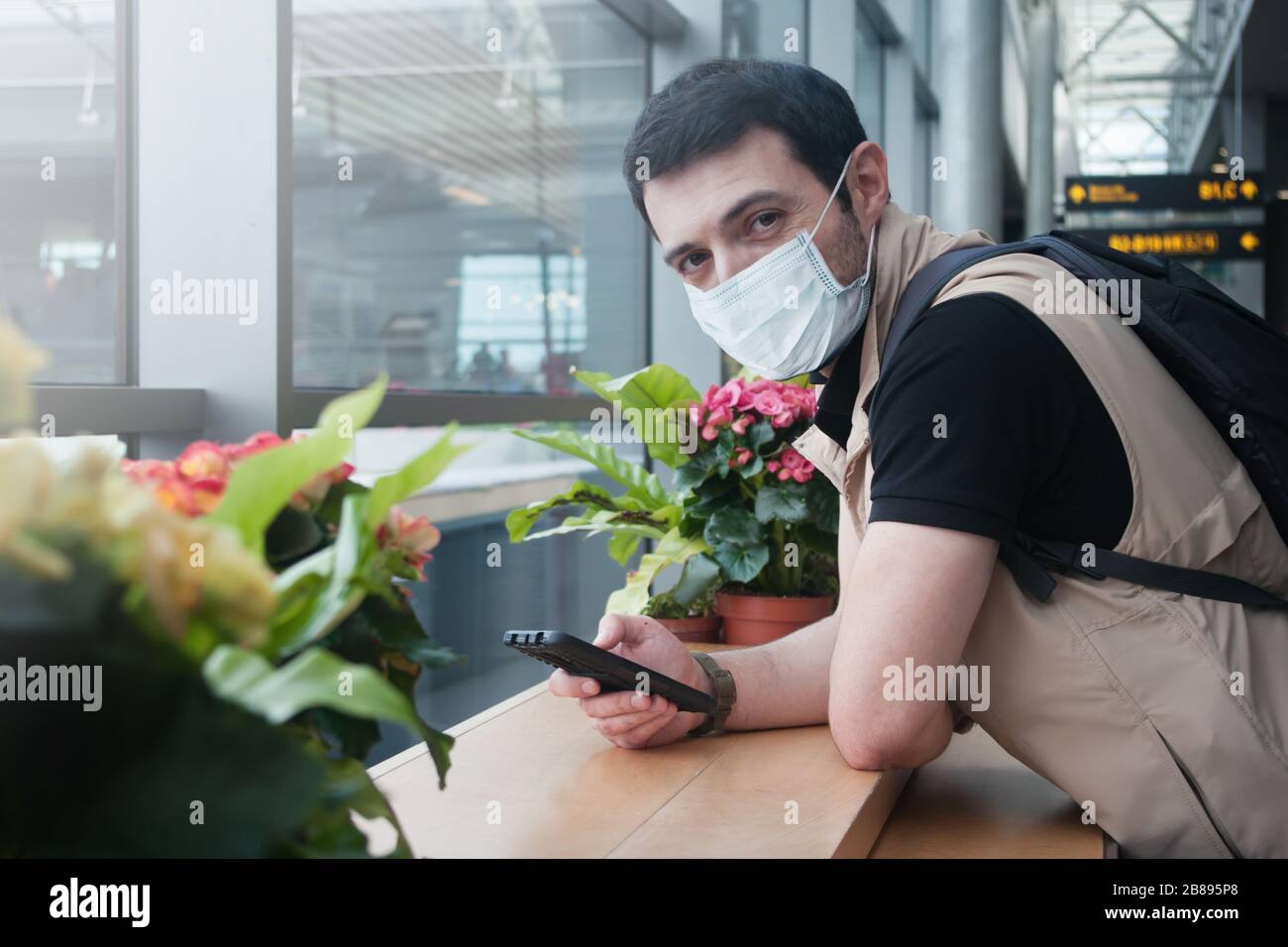 Junger kaukasischer Mann, der eine medizinische Maske trägt und mit seinem Handy entspannt auf sein Licht in der Abflughalle am Flughafen wartet Stockfoto