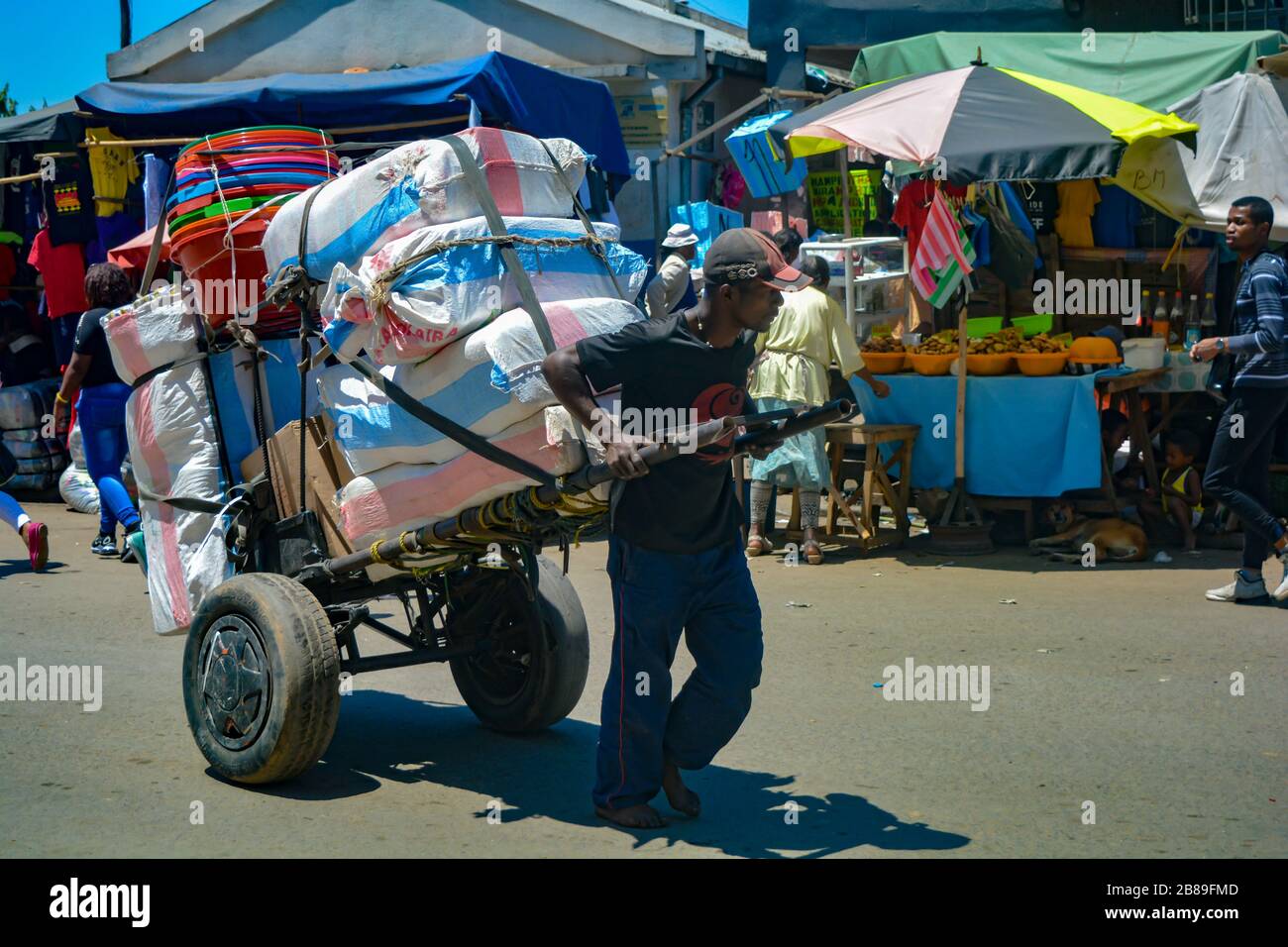 Antananarivo, Madagaskar, Afrika - 01/12/20: Ein schwarzer Mann, barfuß, trägt einen schweren Handwagen voller Beutel mit Mehl und Eimern. Harte Arbeit in einem Markt Stockfoto