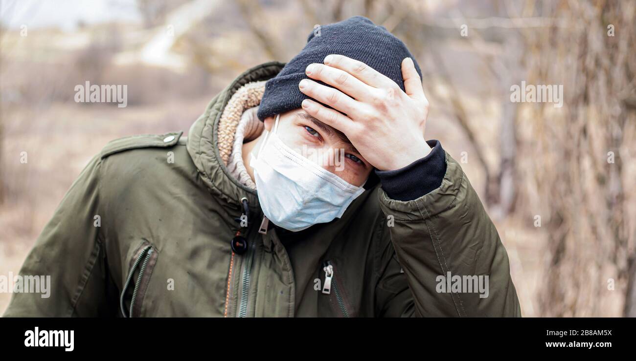 Kerl in einer medizinischen Einwegmaske hält seine Stirn mit der Hand und überprüft die Körpertemperatur. Epidemische Kovid-19 Quarantäne virale Infektion der Atemwege Stockfoto