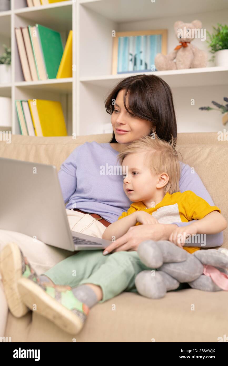 Blonden, haariger Junge, umarmt von einer Mutter, die auf dem Sofa sitzt und neugierige Cartoons auf dem Laptop zu Hause sieht Stockfoto