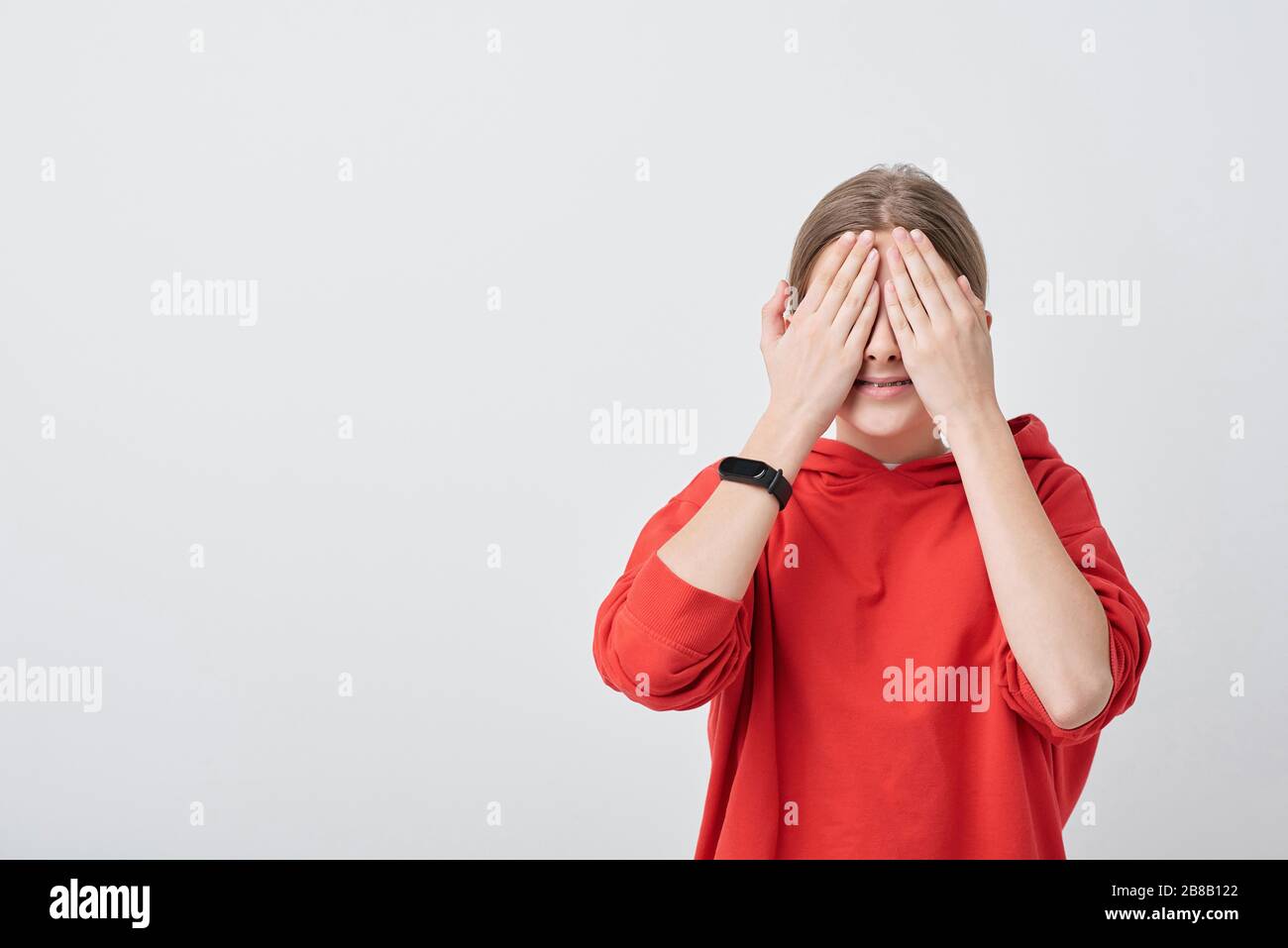 Lächelndes Mädchen im Teenager-Alter in roter Kapuze, das ihre Augen beim Hide-and-Seek Game mit den Händen bedeckt, während sie vor der Kamera steht Stockfoto
