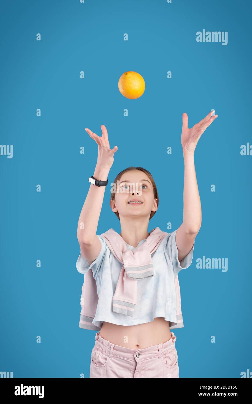 Energiegeladenes Teenager-Mädchen, das beim Spielen vor der Kamera vor blauem Hintergrund isoliert frisches Orange wirft Stockfoto
