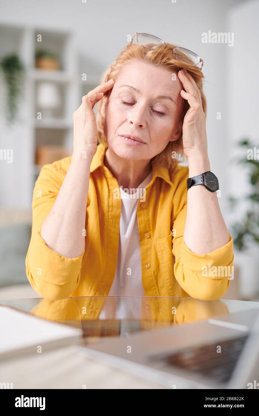 Müde reife Geschäftsfrau in der Casualwear, die den Kopf berührt, während sie am Arbeitsplatz in der häuslichen Umgebung unter Kopfschmerzen leidet Stockfoto