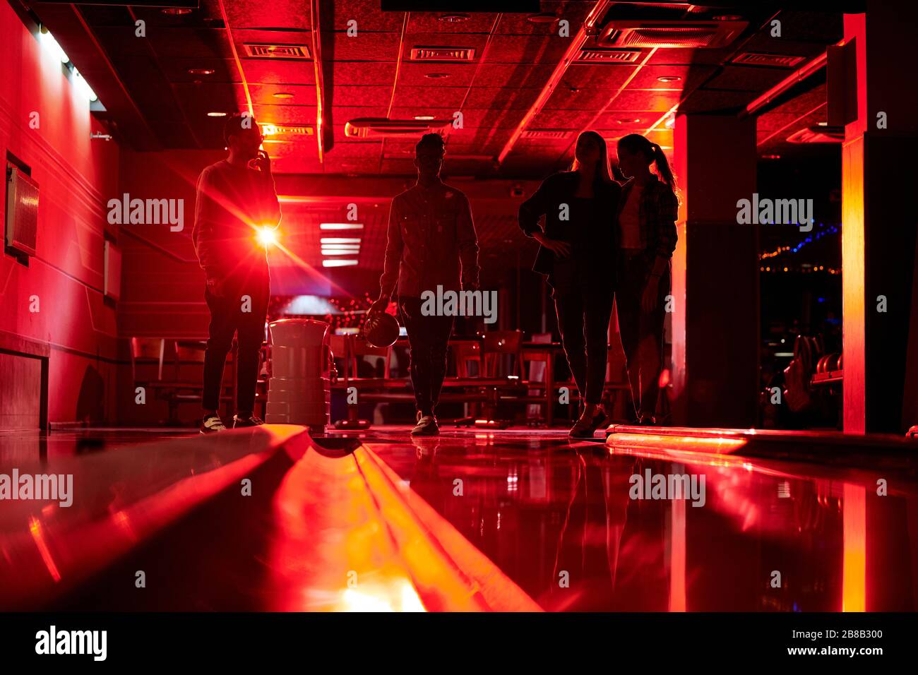 Silhouetten von vier jungen Leuten, die an der Bowlingbahn in dunklem Raum stehen, während einer von Jungs den Ball werfen wird Stockfoto