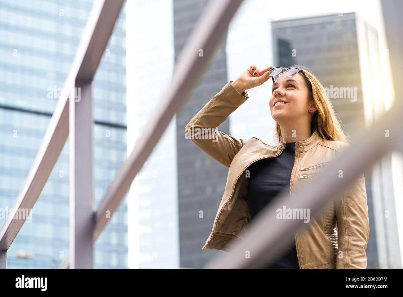 Fröhliche lächelnde Frau in der Großstadt mit Wolkenkratzern. Person, die Sonnenbrille hebt, nach oben schaut und lächelt. Positiver urbaner Lebensstil. Stockfoto
