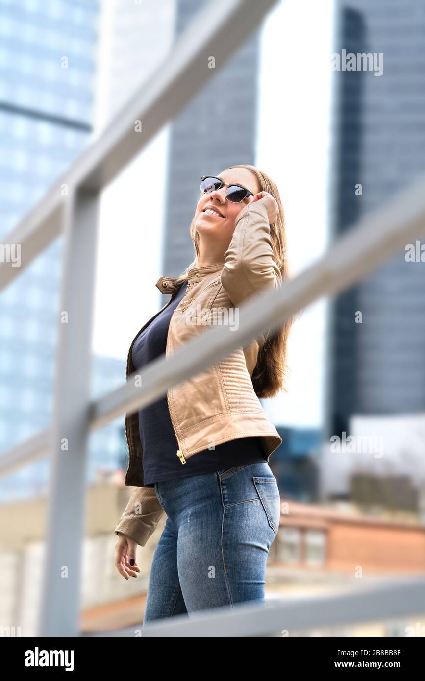 Fröhliche junge Frau in der Großstadt mit Hochhäusern und Wolkenkratzern. Elegante und selbstbewusste Dame mit Sonnenbrille. Vertikaler Schuss. Stockfoto