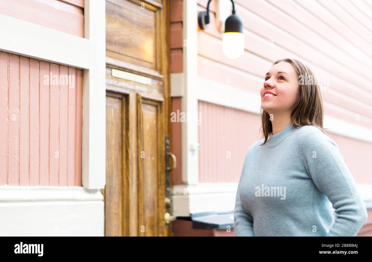 Außenporträt lächelnder Frau mit Pullover. Fröhliche Person, die vor einem alten Gebäude in der Altstadt aufblickt. Holzhaus und Tür in der Stadt. Stockfoto