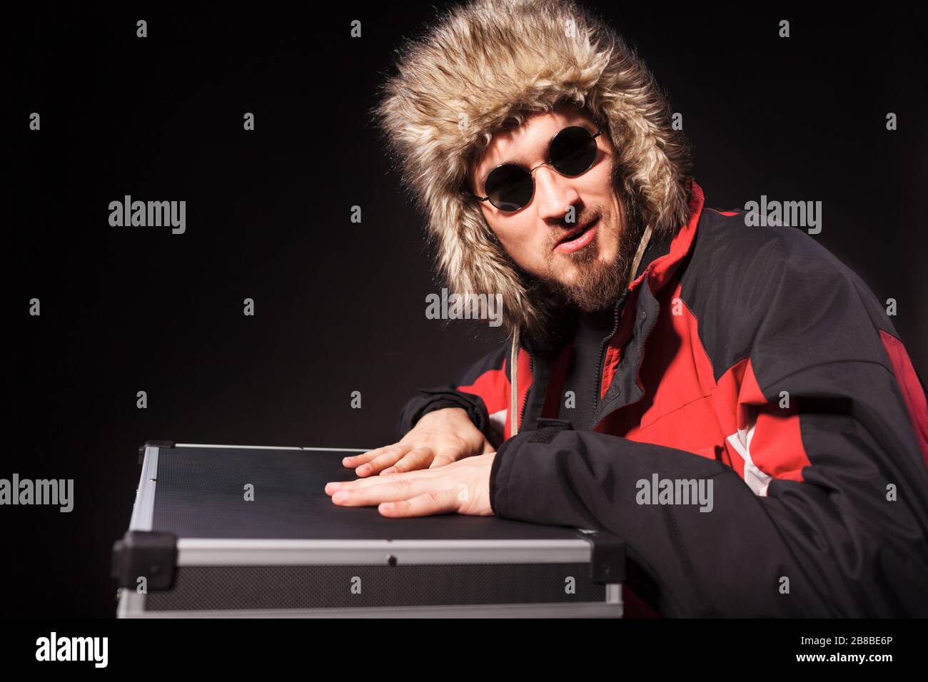 Seitenansicht der gut aussehenden Herren, die auf Runden im Koffer sitzen. Gutaussehender Typ in Sonnenbrille mit Fellhut. Isoliert auf schwarzem Studiohintergrund. Konz Stockfoto