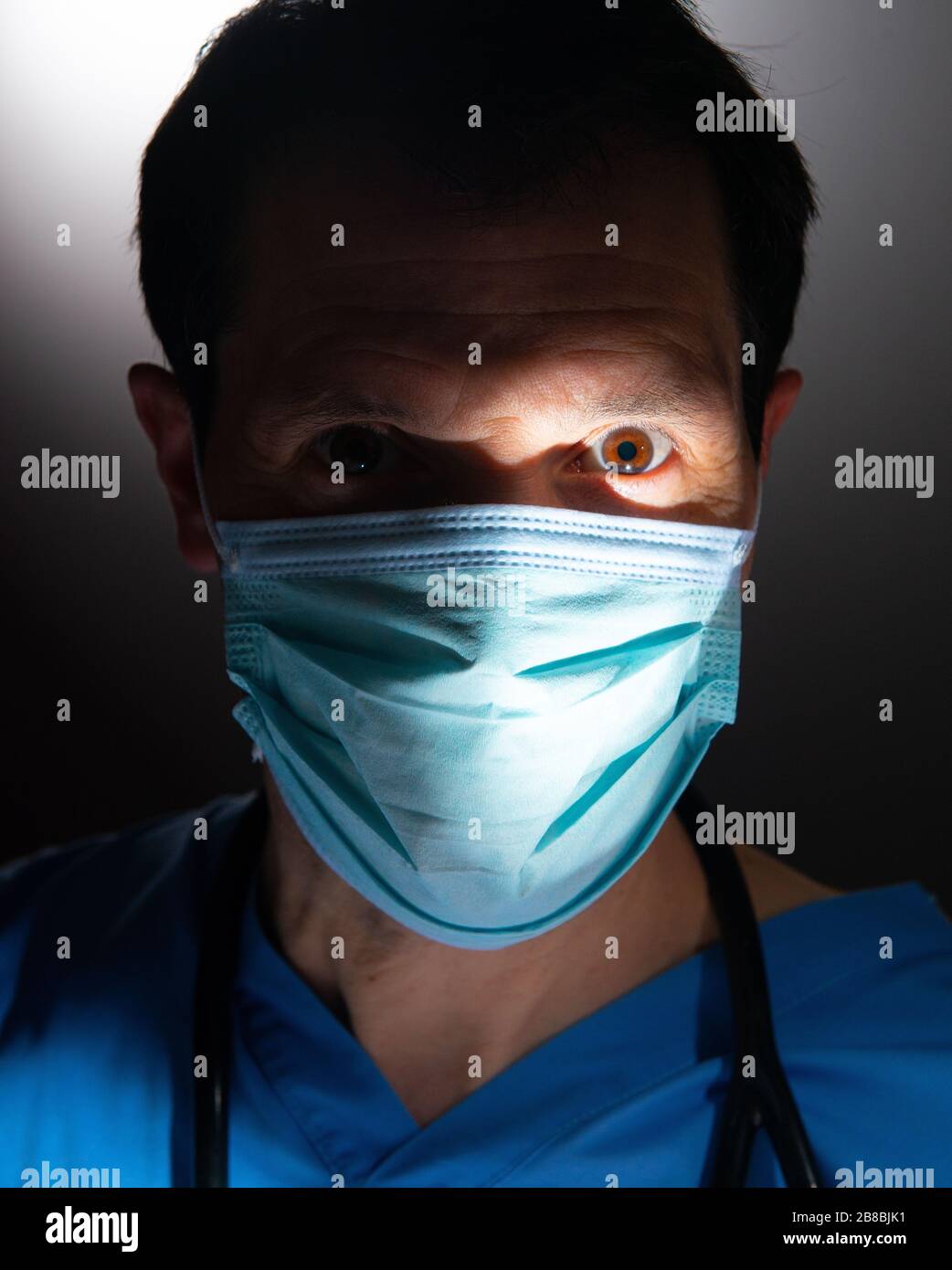 Nahaufnahme eines betroffenen Arztes mit Halbgesicht im Schatten und mit blauen Schruben, mit chirurgischer Gesichtsmaske und Stethoskop, vor dunklem Hintergrund. Stockfoto