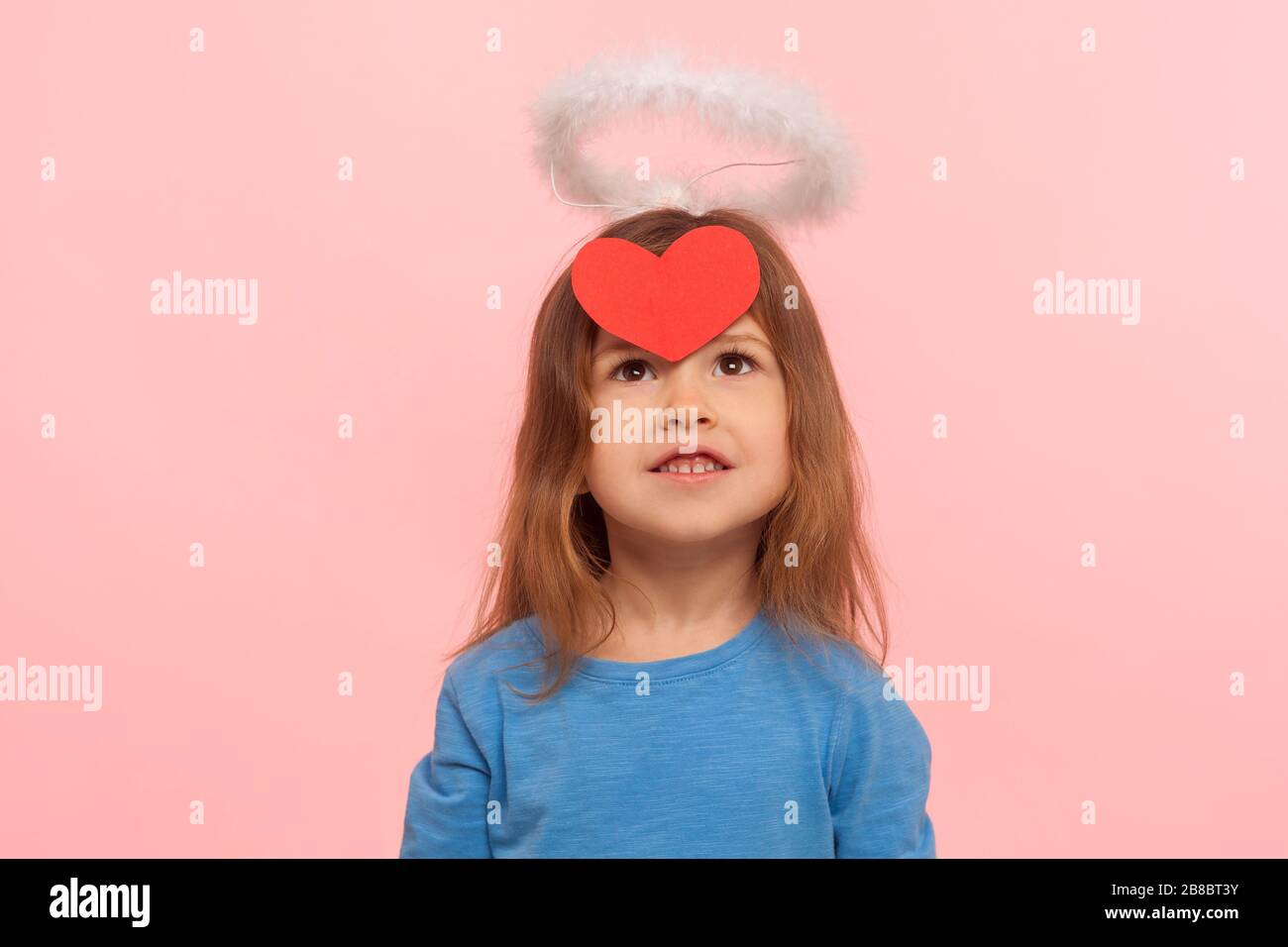 Kleiner kupiger Engel für Valentinstag. Porträt eines träumerisch liebenswerten Vorschulmädchens mit Nimbus, das auf dem roten Papierherzwand auf der Stirn aufblickt. i Stockfoto