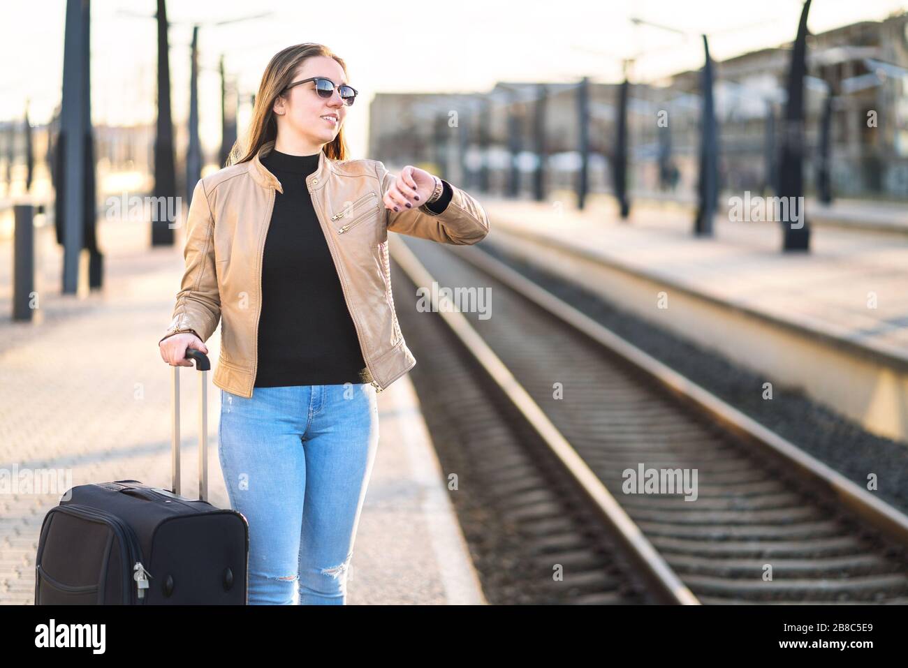 Frau, die sich die Zeit ansieht und zuschaut, während sie auf den Zug wartet. Lächelnde und fröhliche Reisende, die am Bahnhof und am Bahnsteig stehen. Stockfoto