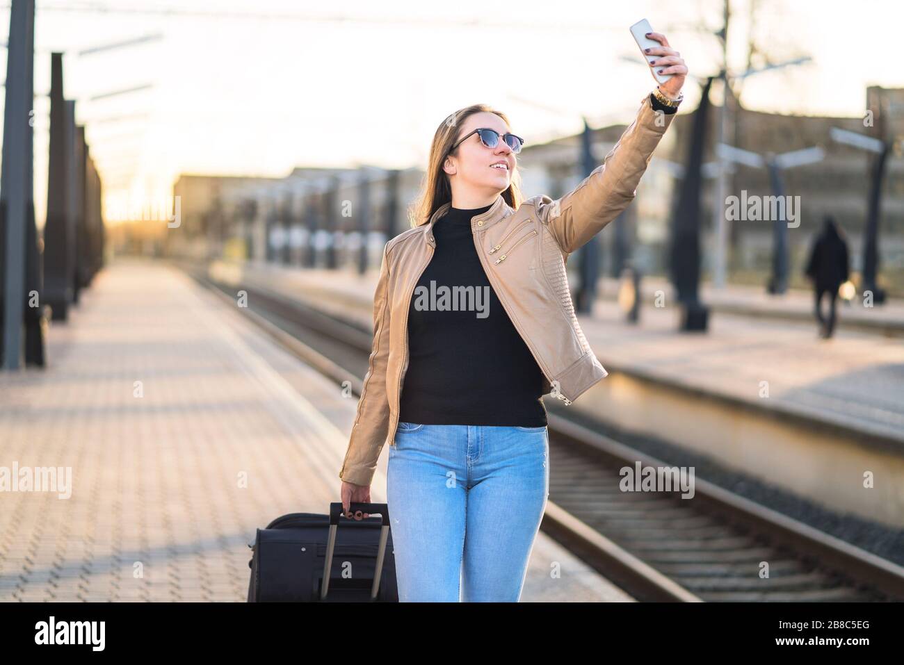 Frau nimmt selfie mit Handy im Bahnsteig am Bahnhof. Lächelnde und fröhliche Dame, die mit Smartphone fotografiert. Stockfoto