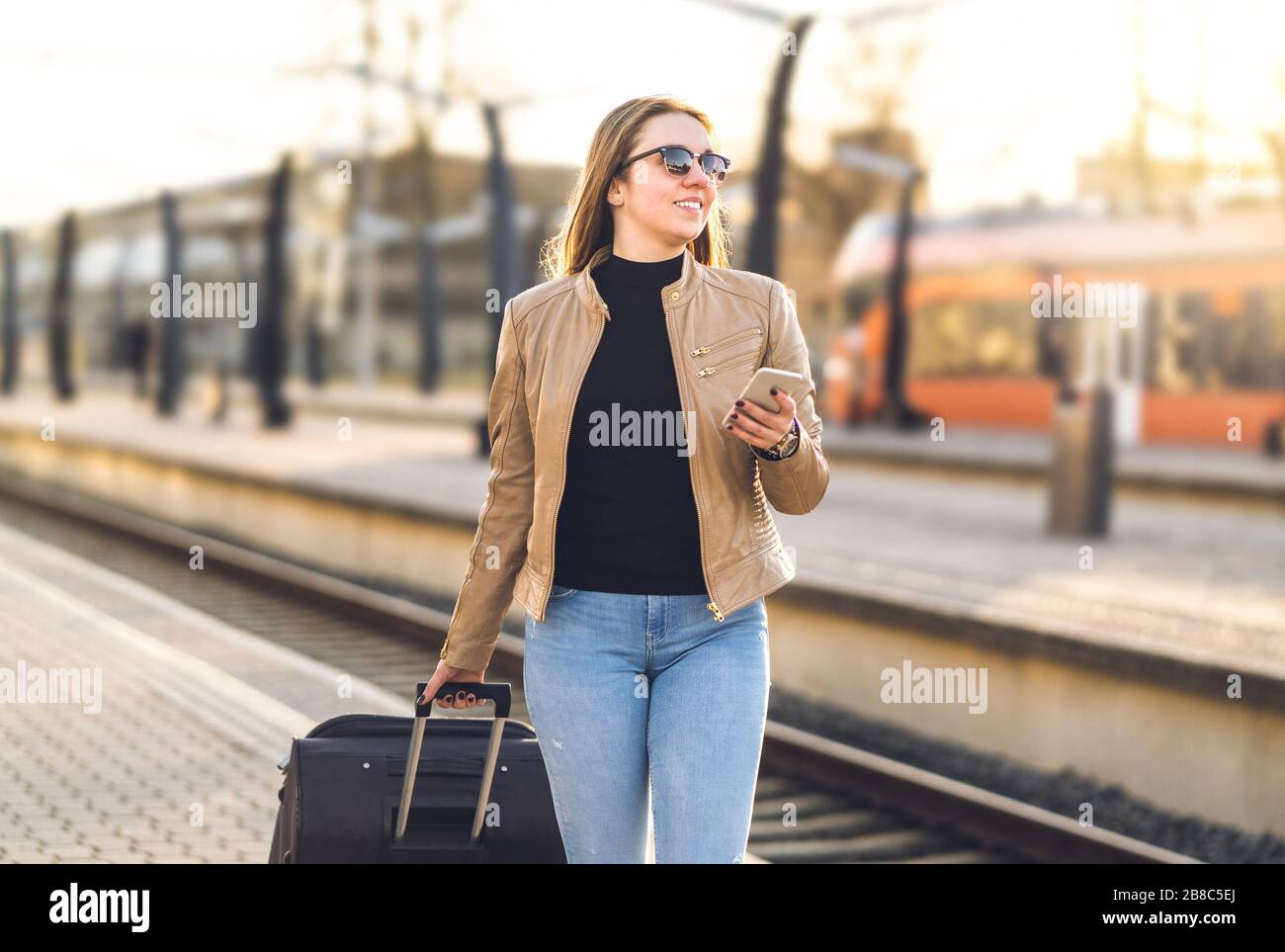 Frau, die mit Smartphone im Bahnhof spazieren geht. Fröhliche Reisende, die Koffer und Gepäck auf der Plattform ziehen und dabei das Handy halten. Stockfoto