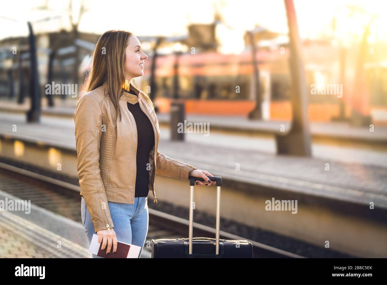 Lächelnde Frau mit Gepäck im Bahnhof. Fröhliche Dame, die mit Koffer, Pass und Ticket am Bahnsteig steht. Eisenbahn-Reise- und Lifestyle-Konzept. Stockfoto