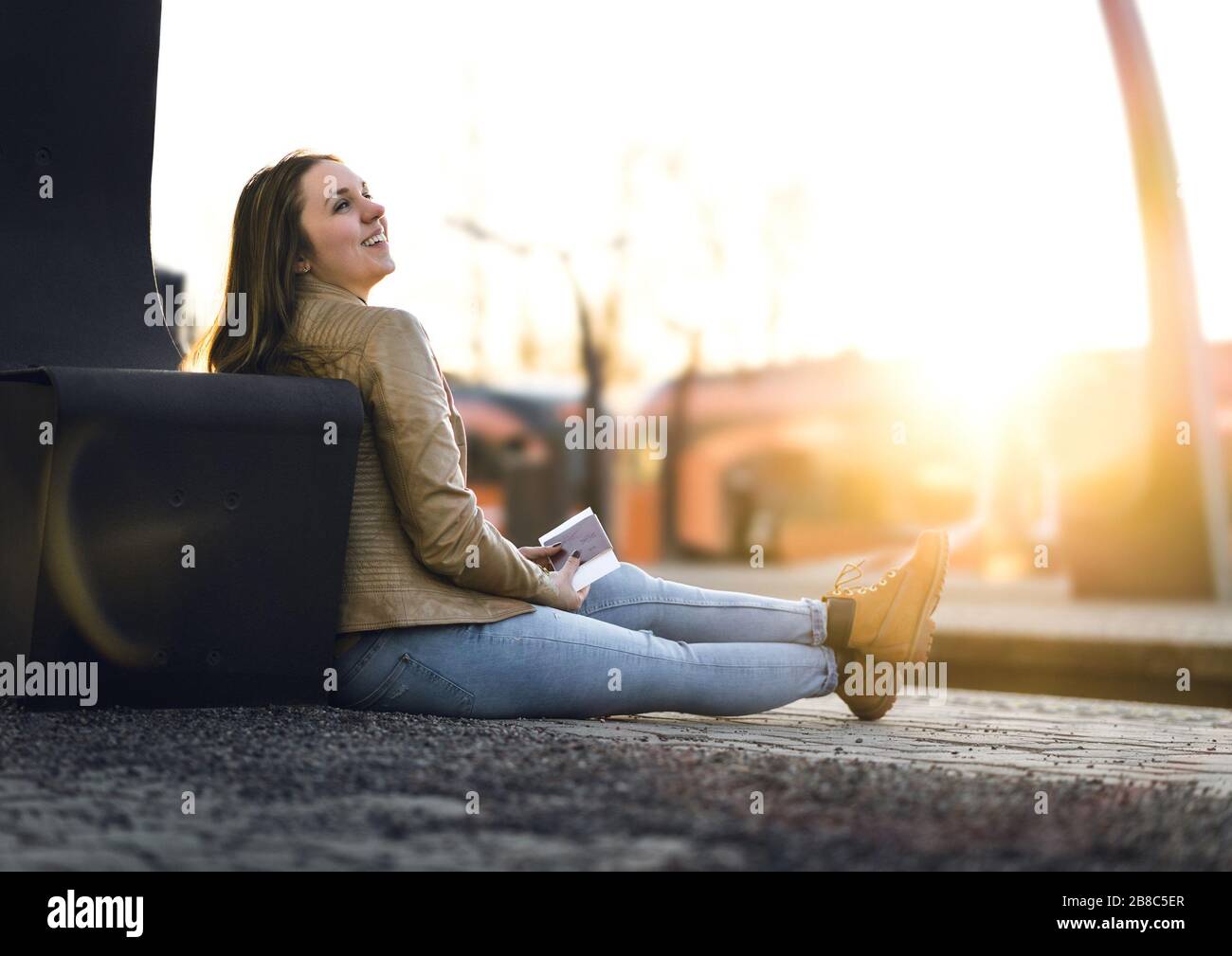 Lachende und fröhliche Frau wartet am Bahnhof. Mit Pass und Ticket am Boden sitzen. Urlaub machen und reisen. Stockfoto