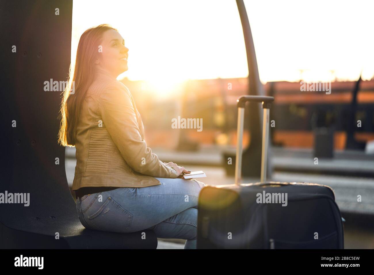 Glückliche Frau bei Sonnenuntergang wartet auf Zug. Dame sitzt mit Gepäck, Gepäck und Koffer auf der Plattform am Bahnhof. Reise- und Lifestyle-Konzept. Stockfoto
