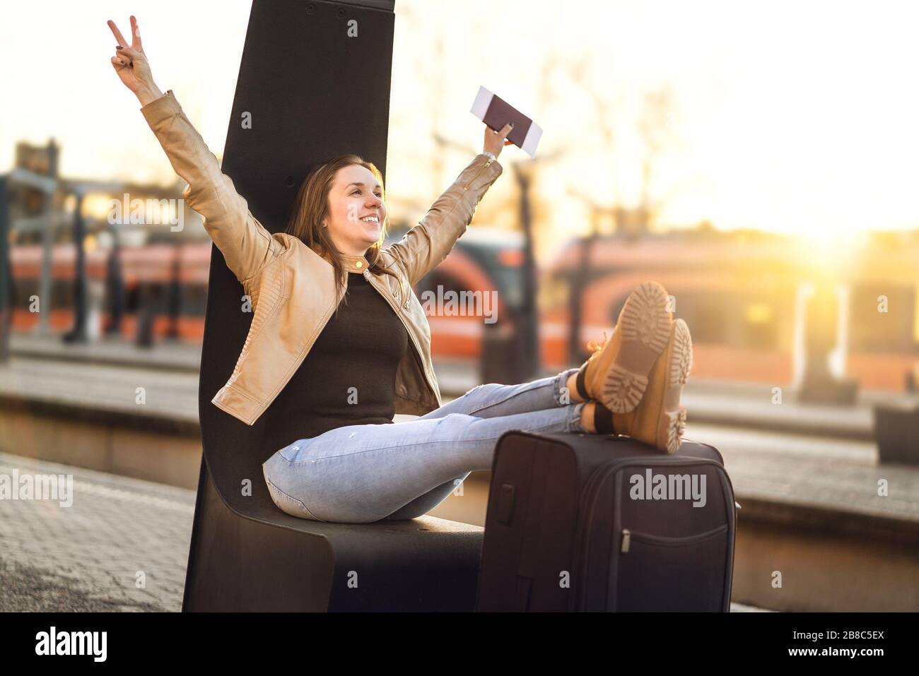 Endlich im Urlaub. Frau feiert am Bahnhof, um auf lustige Reise zu gehen. Fröhlicher lächelnder Mensch mit Beinen und Schuhen auf Koffer und Gepäck. Stockfoto