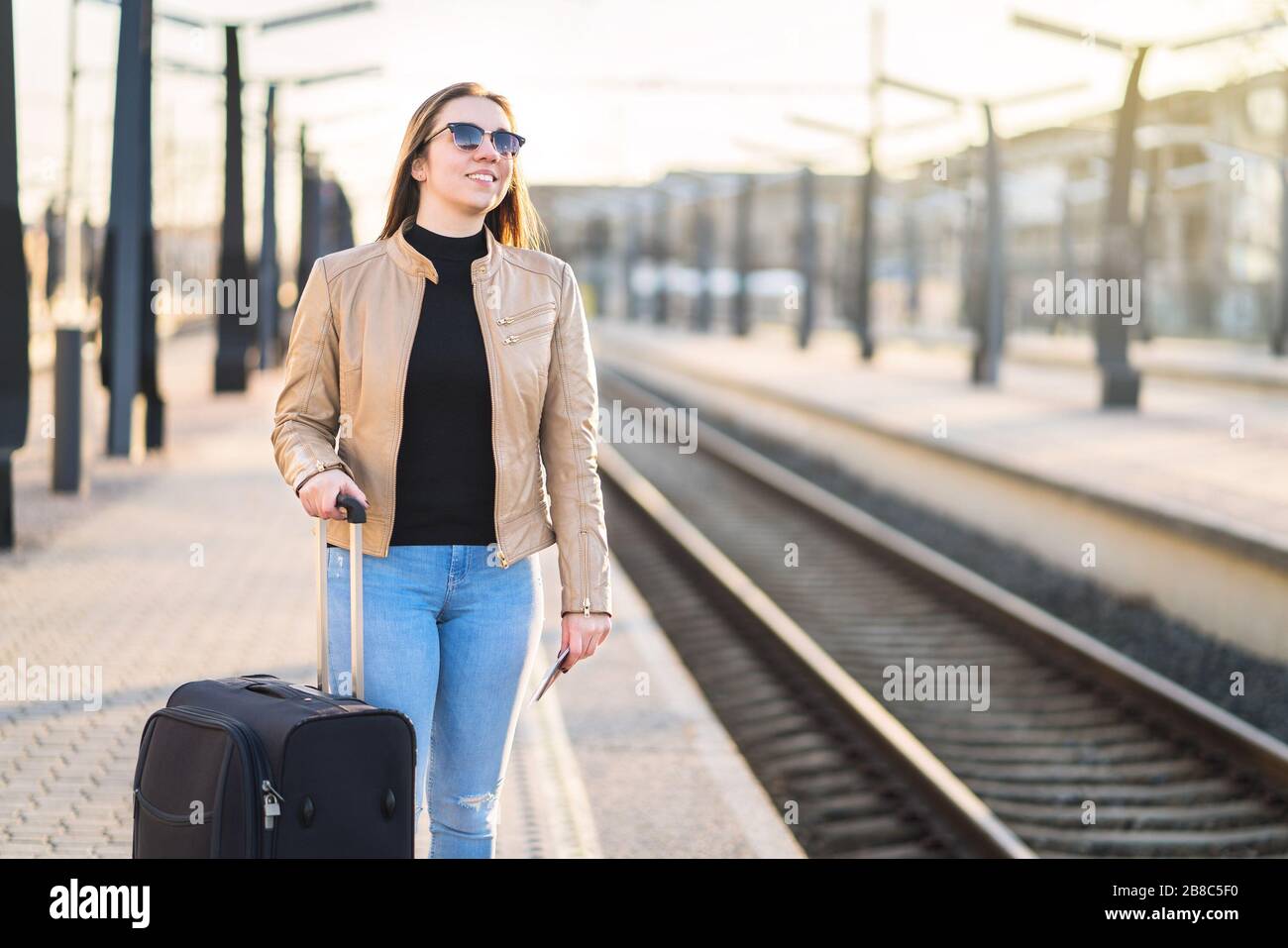 Frau wartet am Bahnhof auf den Zug. Fröhliche lächelnde Dame am Bahnsteig, die auf Bahnfahrt geht. Person mit Pass und Ticket und Blick auf die Spuren. Stockfoto