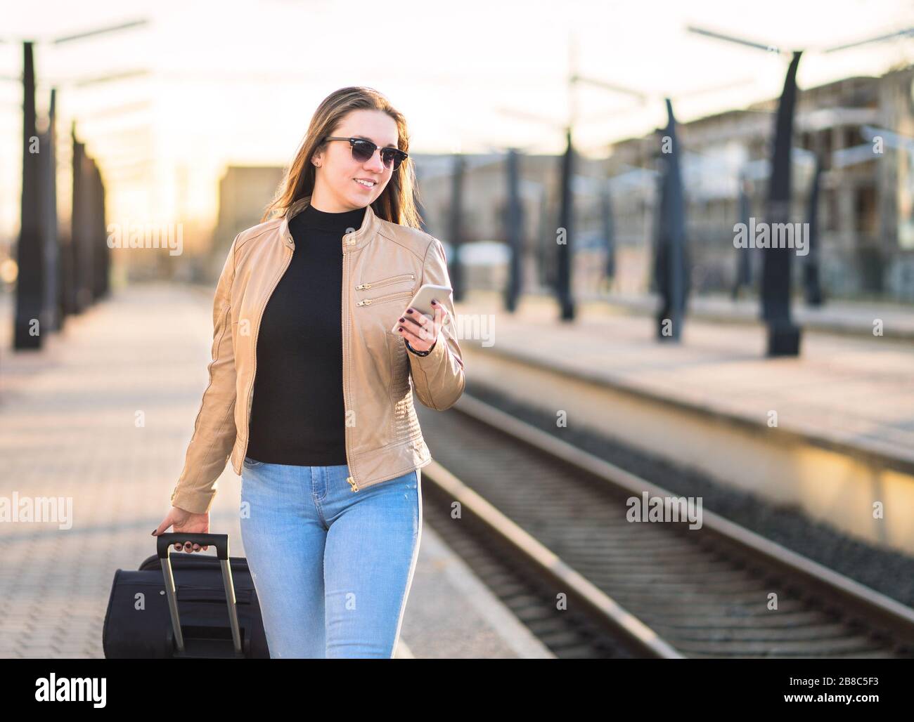 Frau, die im Bahnhof spazieren geht und Smartphone benutzt. Passagier kauft elektronische digitale Tickets mit Handy. Glückliche Dame zieht Koffer- Stockfoto