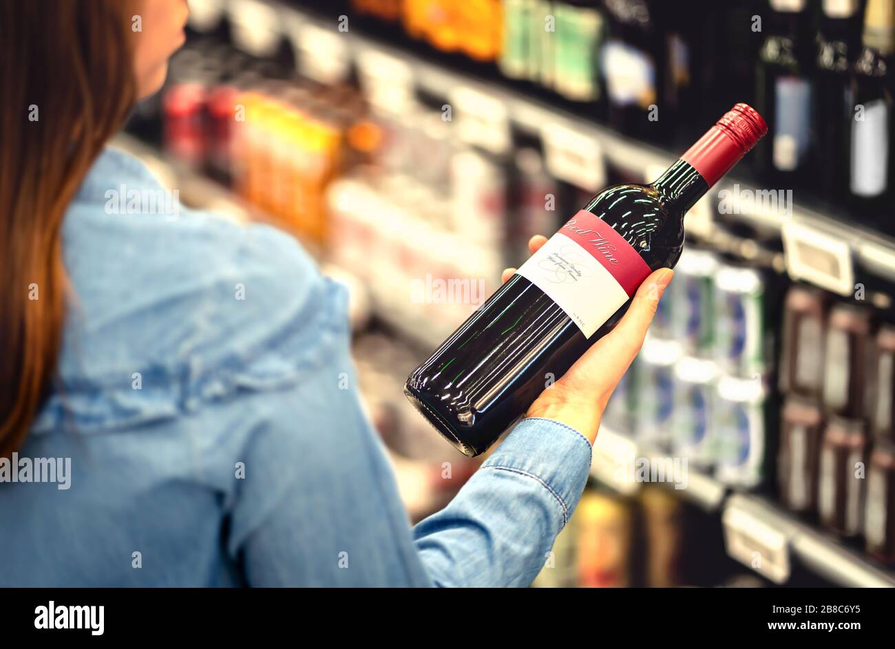 Frau, die das Etikett der Rotweinflasche im Spirituosenladen oder Alkoholbereich im Supermarkt liest. Regal voller alkoholischer Getränke. Stockfoto