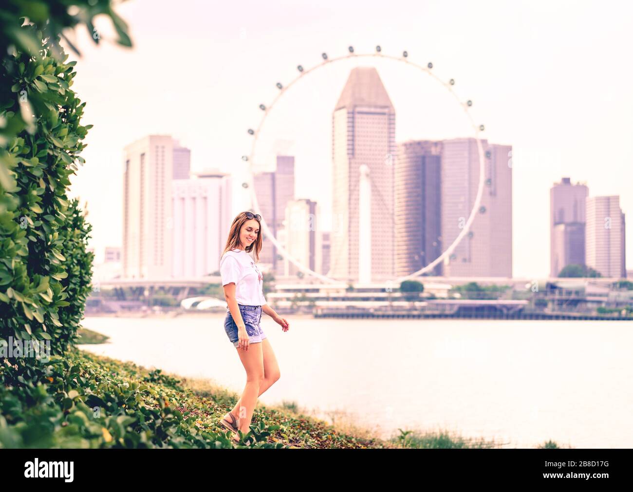 Positive trendige Frau in Jeans-Shorts, die in einem Park im Freien spazieren gehen und die Skyline der Stadt im Hintergrund sehen. Fröhlicher modischer Lebensstil. Stockfoto