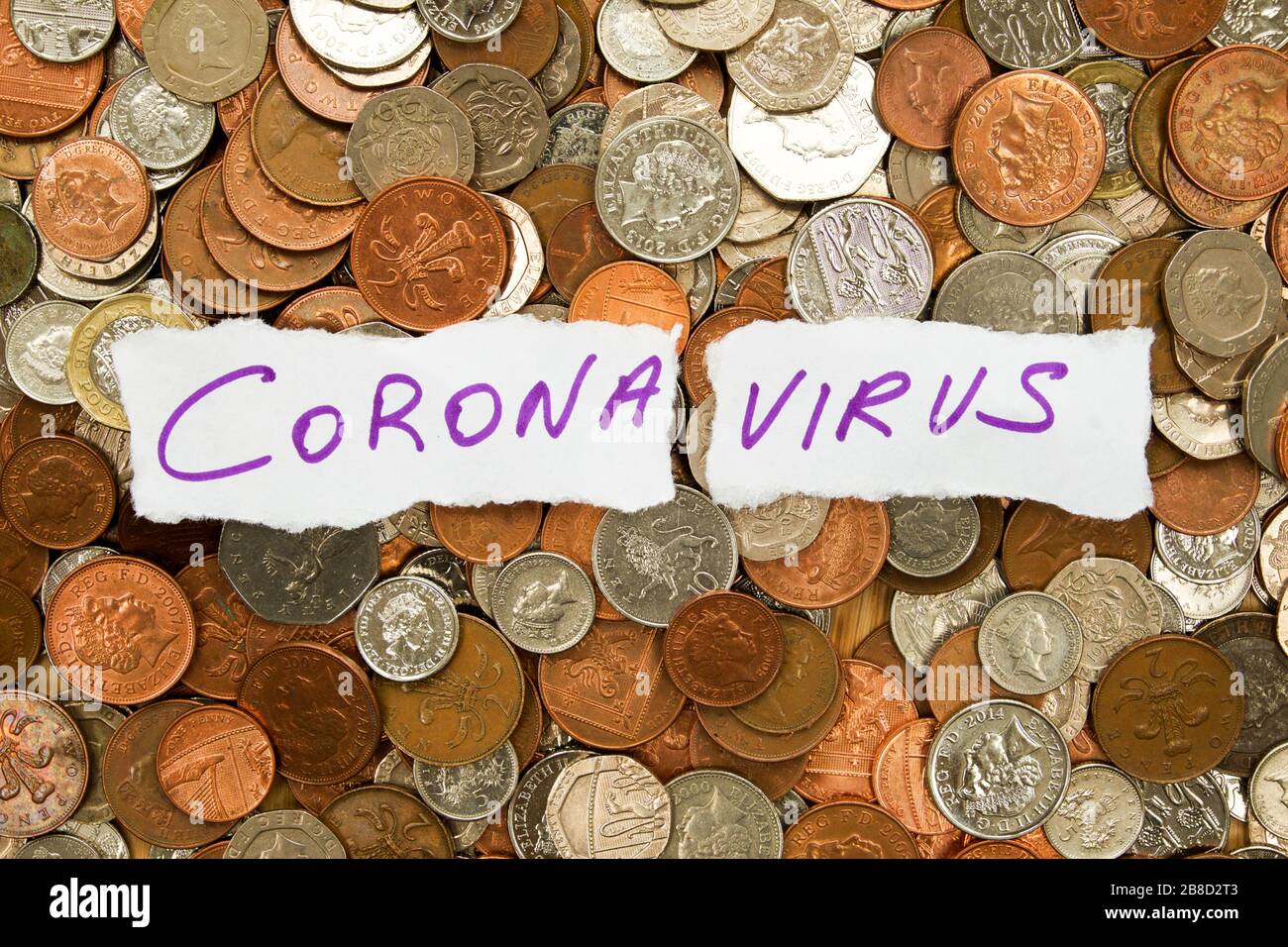 Die Wörter Corona-Virus in lila Tinte auf zwei Teilen zerrippten weißen Papiers, das auf Hunderte von Silber- und kupferfarbenen Münzen aufliegt, Pfund Stockfoto
