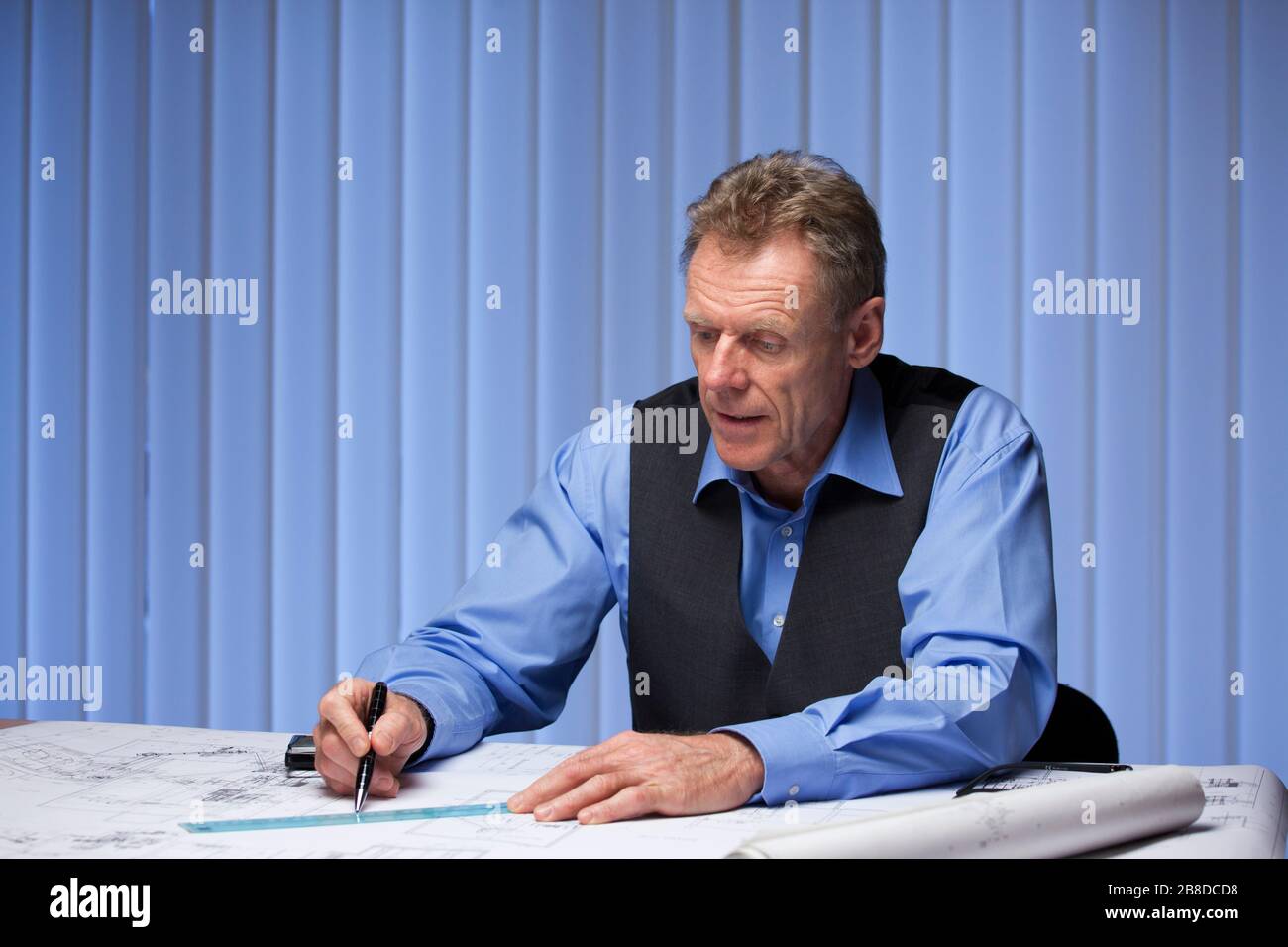 Reifer Architekt oder Ingenieur, der an einem Schreibtisch mit einem Plan mit blauem Bürohintergrund sitzt Stockfoto
