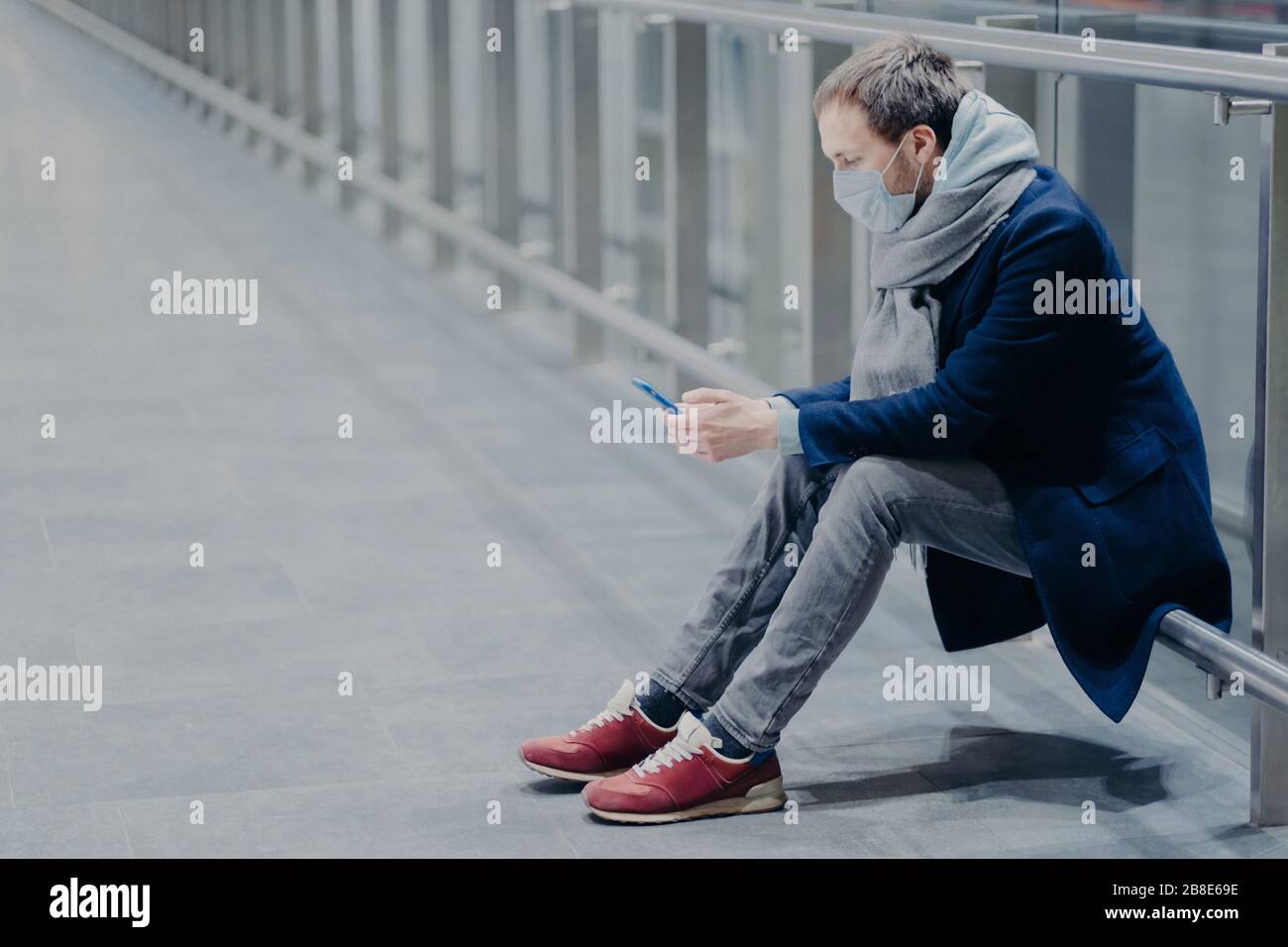 Müde Touristen machen Pause, benutzt Handy, gekleidet in Jacke, Jeans und Turnschuhe, trägt Schutzmaske gegen Coronavirus, kümmert sich um die Gesundheit Stockfoto