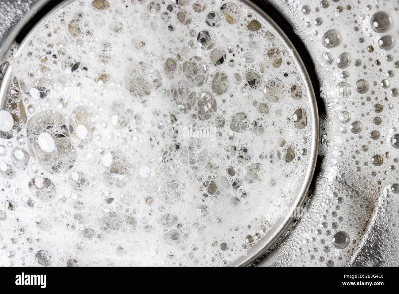 Weißer Schaum mit Reinigungsblasen in einem Waschbecken, Makroansicht. Ablassöffnung mit Seifenblasen im Metallwaschbecken. Mechanisch verstellbare Ablassschraube in der Nähe. Stockfoto