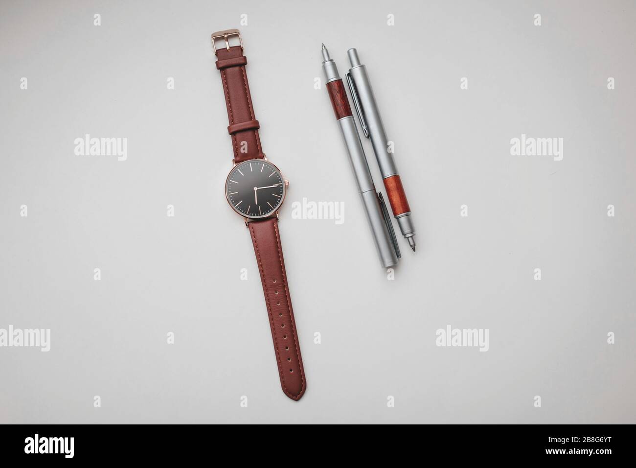 Top View mit schwarz-brauner Armbanduhr und zwei braunen und silbernen Stiften in weißer Kulisse. Stillleben und Bürobedarf Fotografie. Stockfoto