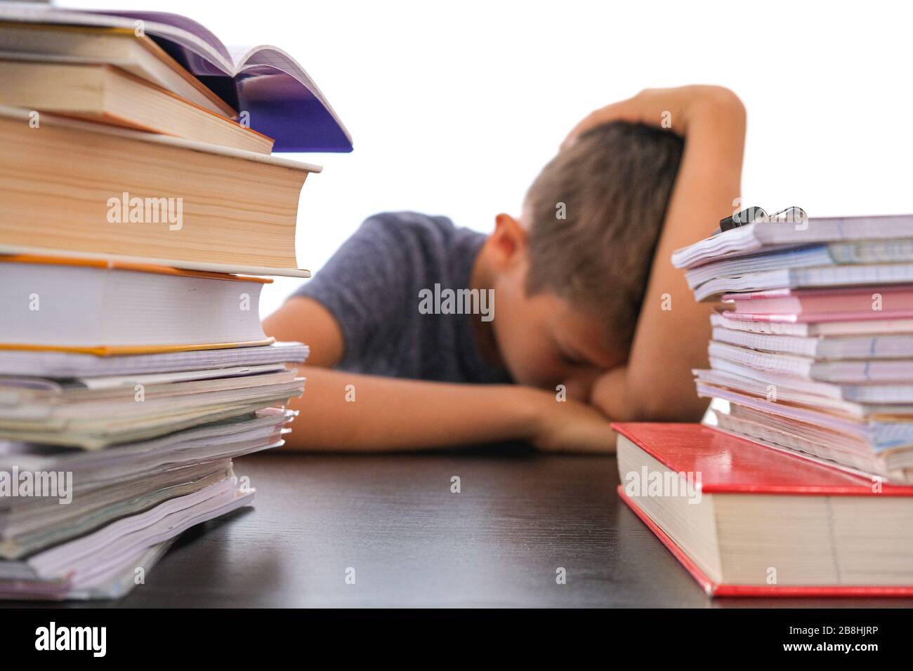 Hefe von Büchern für Schulaufgaben und müde enttäuschte Schuljunge, der seinen Kopf an einem Schreibtisch sitzend senkt Stockfoto