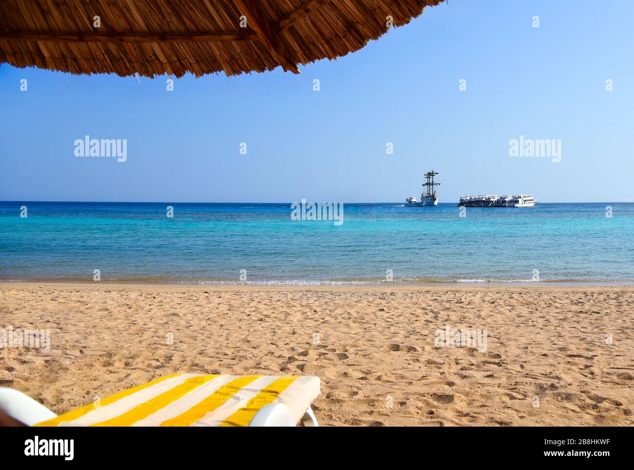 Strandliege unter einem Schilfschirm vor dem blauen Meer mit einer Yacht. Stockfoto