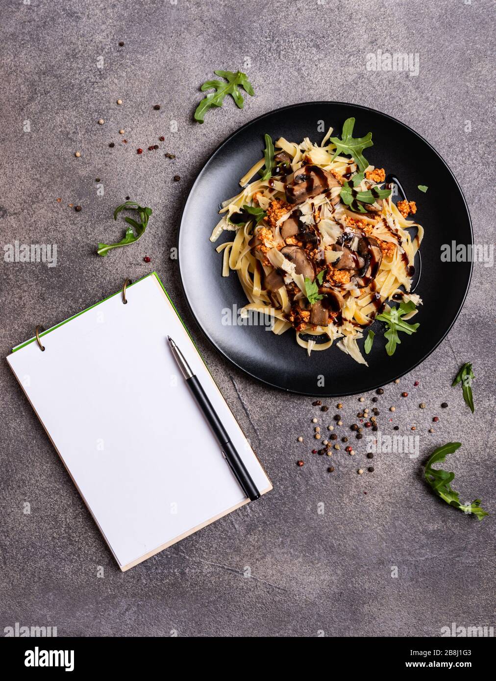 Bolognese Pasta mit Pilzen und Arugula auf schwarzem Teller gebuste Balsamico-Sauce. Ein Buch zum Schreiben von Rezepten liegt auf einem Tisch. Stockfoto