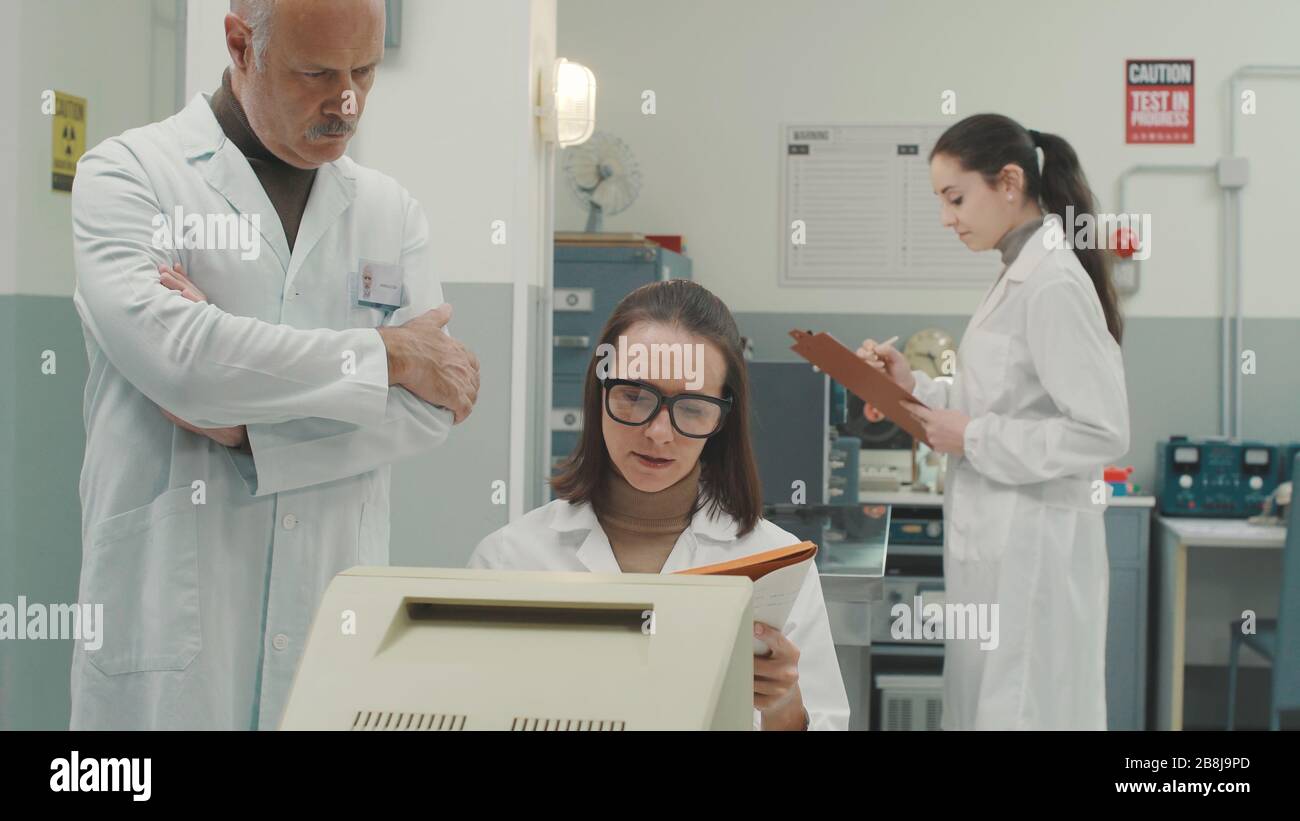 Wissenschaftliches Team, das im Labor mit alten Maschinen und Computern arbeitet, überprüft der leitende Wissenschaftler die Arbeit eines Forschers Stockfoto