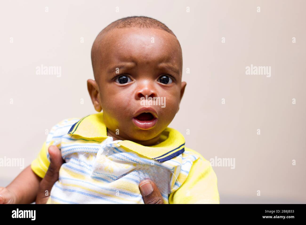 Nahaufnahme des Babys in den Armen seines Vaters, der zu Hause auf der Couch sitzt und dabei die Kamera überrascht beobachtet Stockfoto