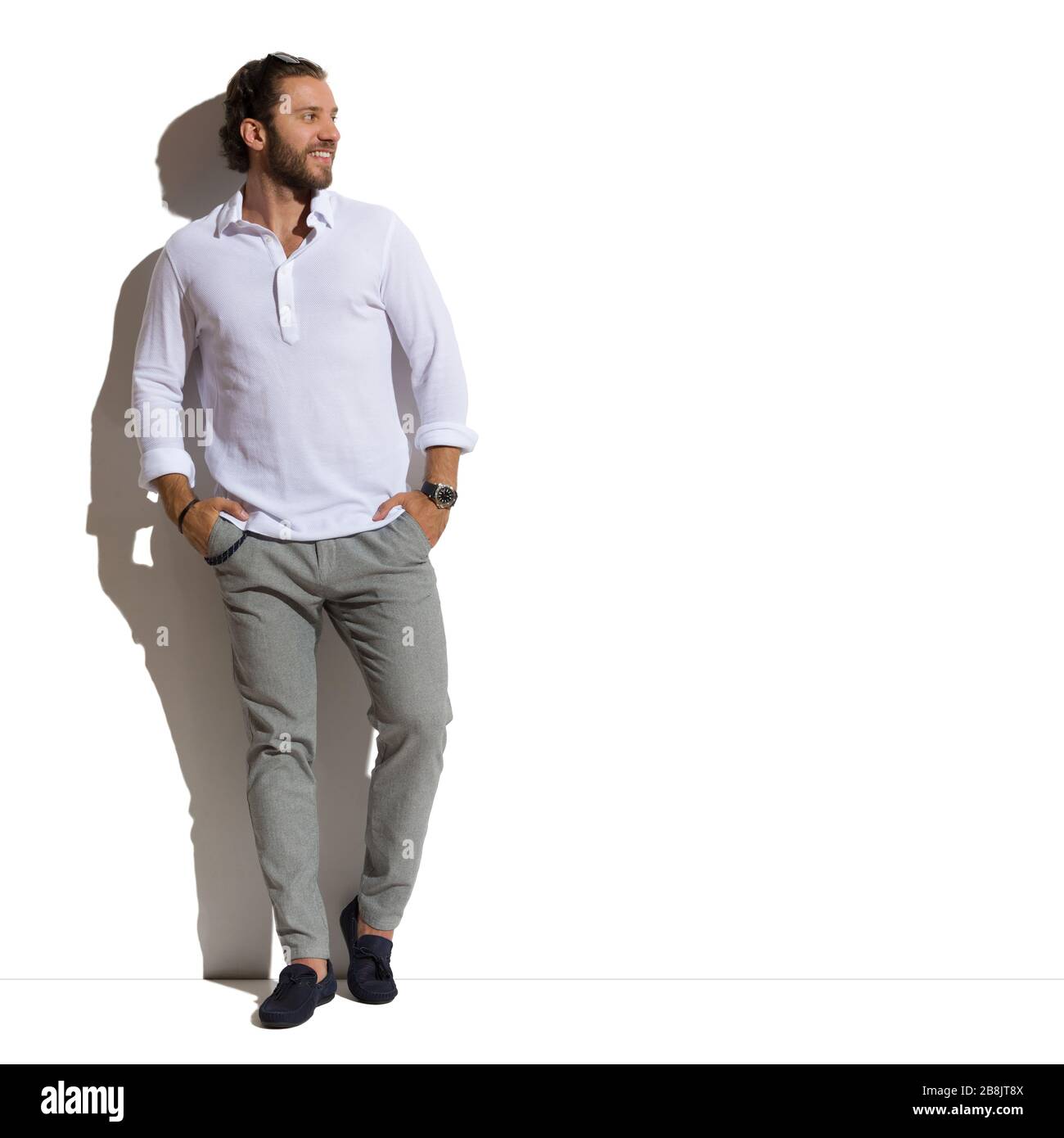 Entspannter, gutaussehender Mann in Mokassins, grauen Hosen und weißem Hemd  steht gegen sonnige Wand mit Händen in Taschen und Blick auf die Seite.  Voll l Stockfotografie - Alamy
