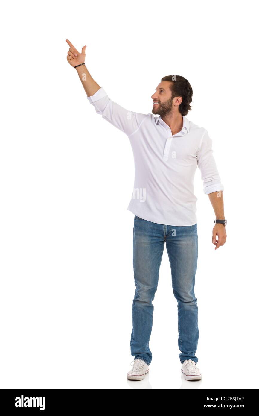 Der junge Mann in Jeans, Turnschuhen und weißem Hemd steht mit aufgehobenem Arm, Blick weg und zeigt nach oben. Vorderansicht. Studio voller Länge isoliert o Stockfoto