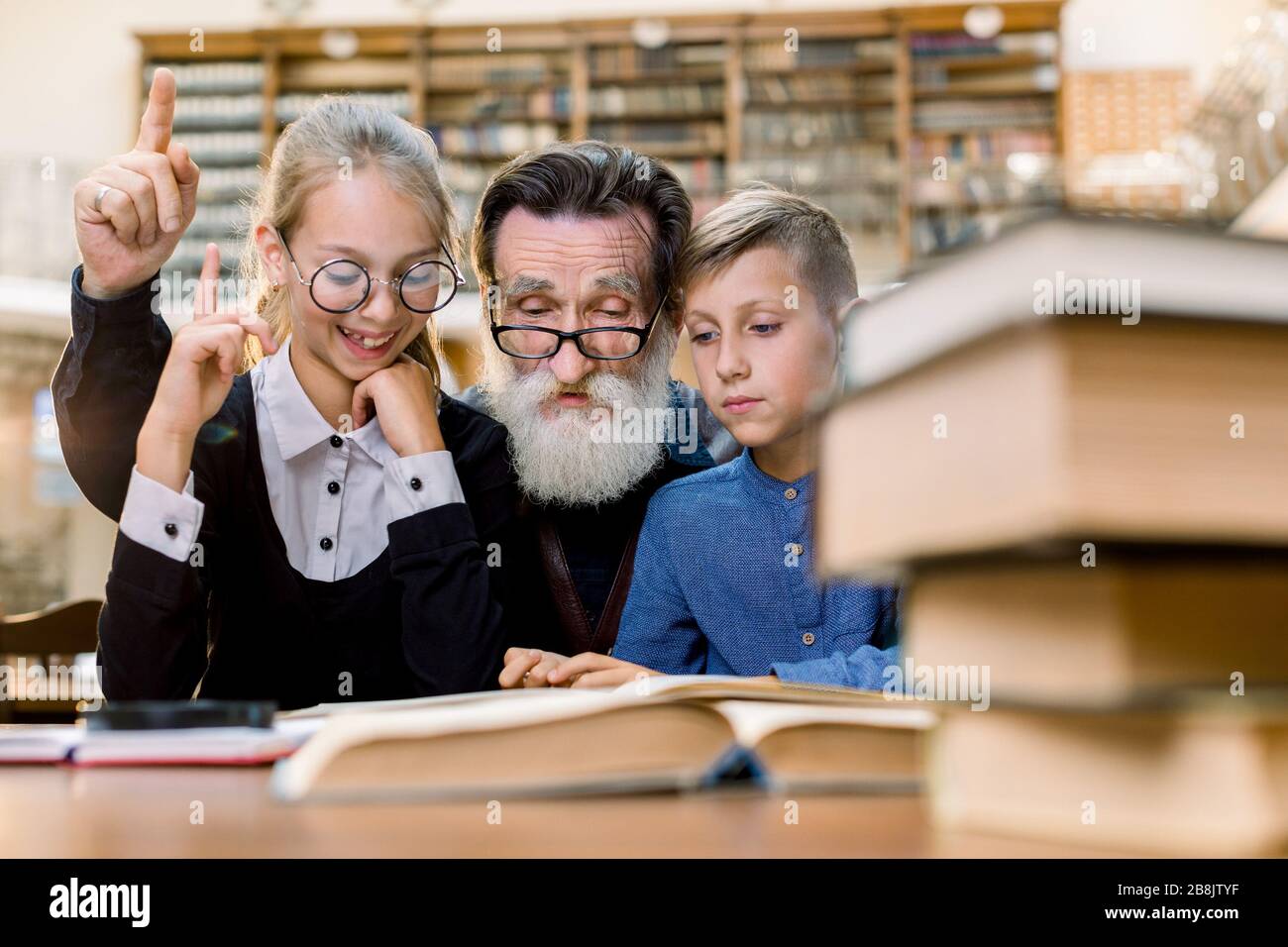 Fröhlich lächelnder Großvater liest Buch mit Enkel und Enkelin, sitzt am Tisch in alter Vintage-Bibliothek. Älterer Mann und das Mädchen sind es Stockfoto