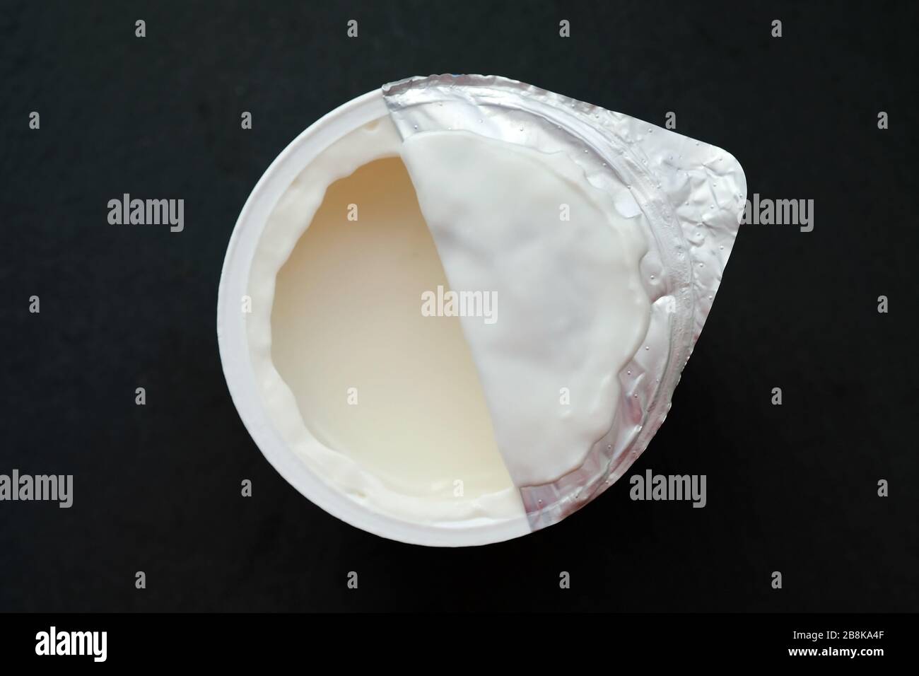 Weißer Joghurt in einem Plastikbecher vor schwarzem Hintergrund. Kinderjoghurt ist von oben halboffen. Stockfoto