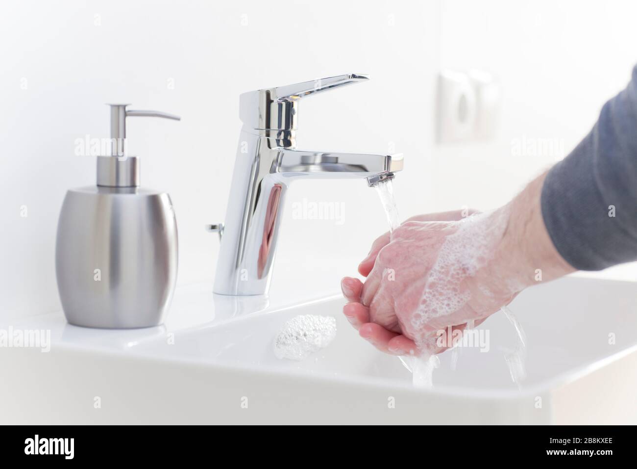 Der Mann wascht seine Hände mit flüssiger Seife in einem weißen Badezimmer, um sich vor Infektionen zu schützen - selektiver Fokus auf den Hahn Stockfoto