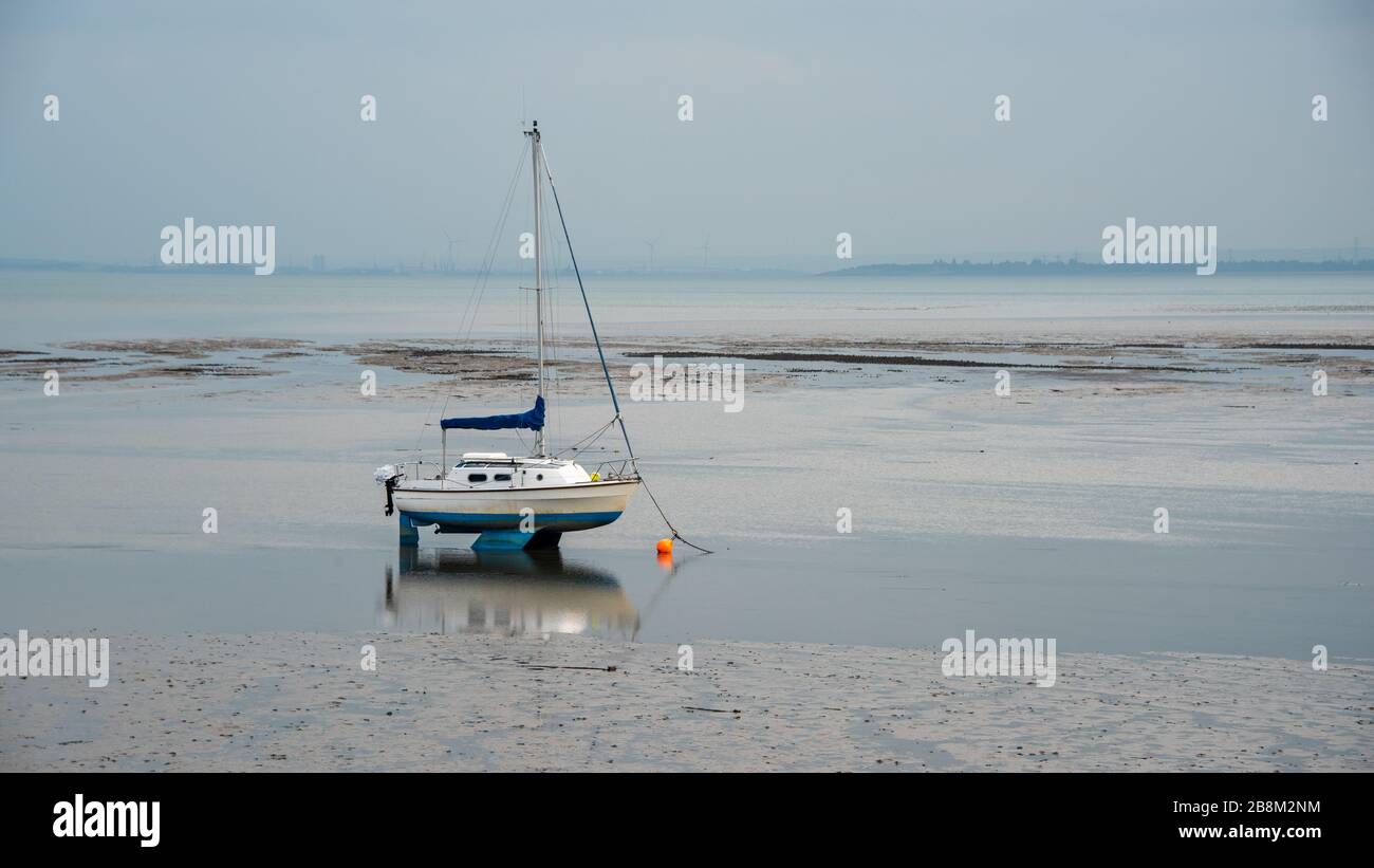 Ein kleines Segelboot auf dem Sand, während die Flut aus dem Wasser gegangen ist und es hoch und trocken gelassen hat. Im nassen Sand gibt es eine Reflexion Stockfoto