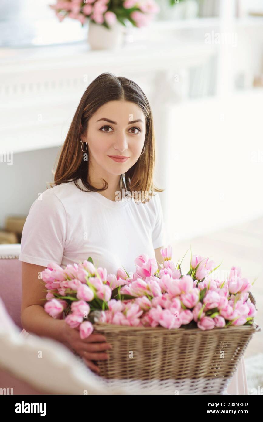 Schöne junge Frau, die auf dem Sofa sitzt und einen Korb mit rosafarbenen Tulpen hat. Stockfoto