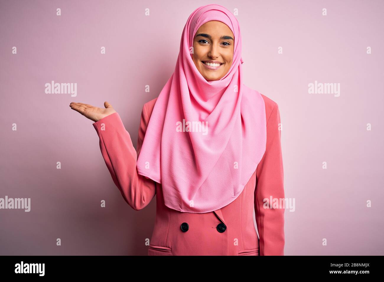 Junge, schöne Brunette-Geschäftsfrau mit pinkfarbenem muslimischem Hijab und Geschäftsjacke lächelnd fröhliche Darstellung und zeigen mit Handfläche Lookin Stockfoto