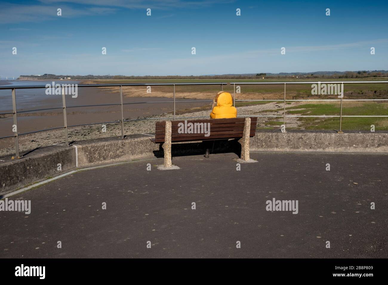 Eine einsame Figur in einer gelben Jacke, die auf einer Bank mit Blick auf die Severn-Estuary und die Severn-Brücke sitzt Stockfoto