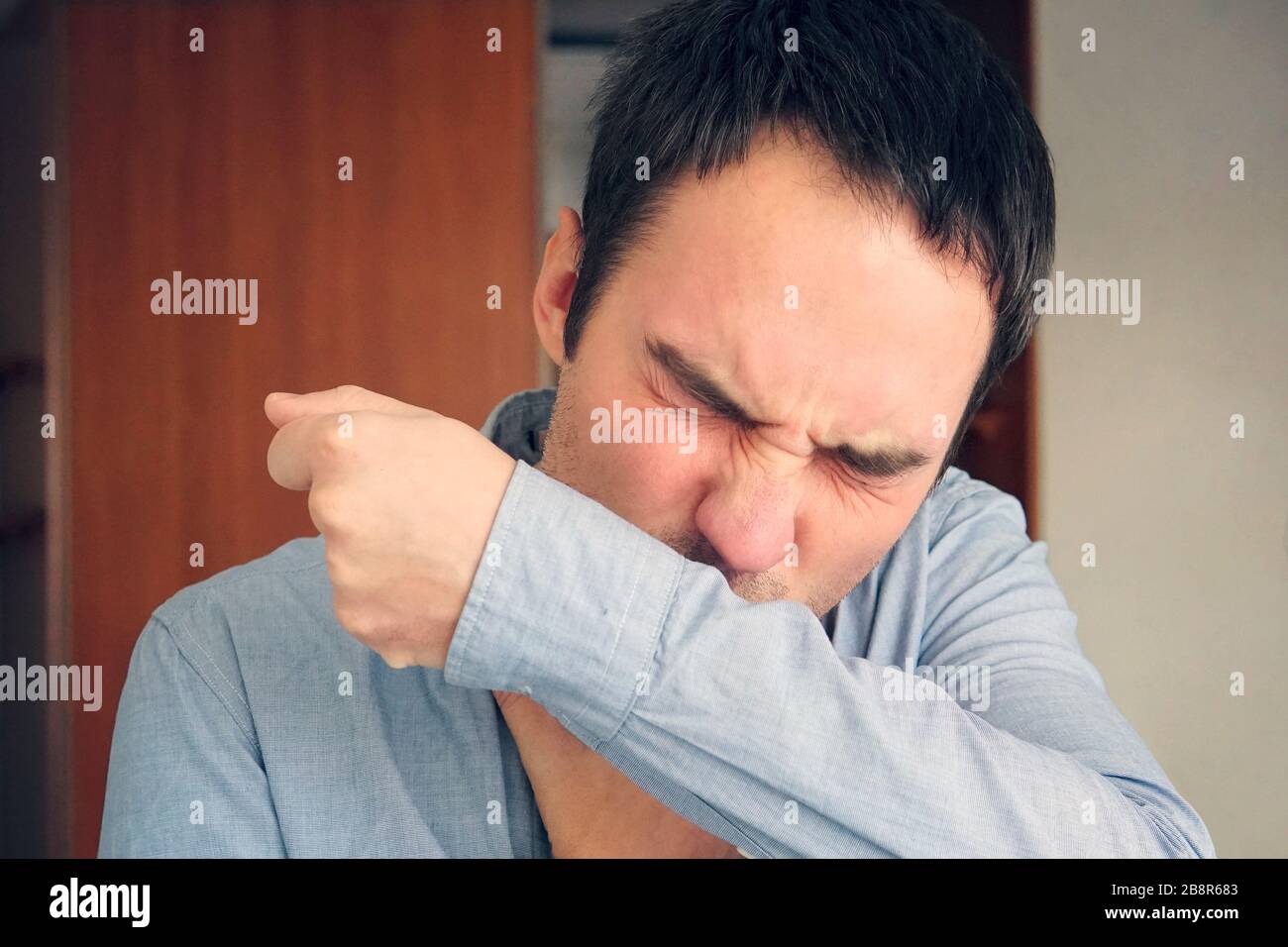 Mann, der die Grippe hat und auf dem Ärmel im Krämer ihres Arms niest. Ein junger Kerl hustet in den Stoff seines Hemdes, um die Verbreitung von Viren zu verhindern Stockfoto