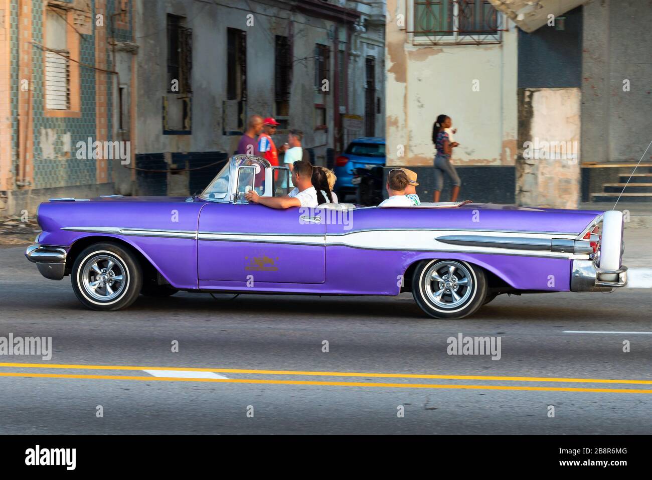 Oldtimer mit Touristen, die vor Konstruktionen in schlechtem Zustand in der Malecon-Promenade in Havanna, Kuba, vorbeifahren. Vehikel in leuchtenden Farben Stockfoto