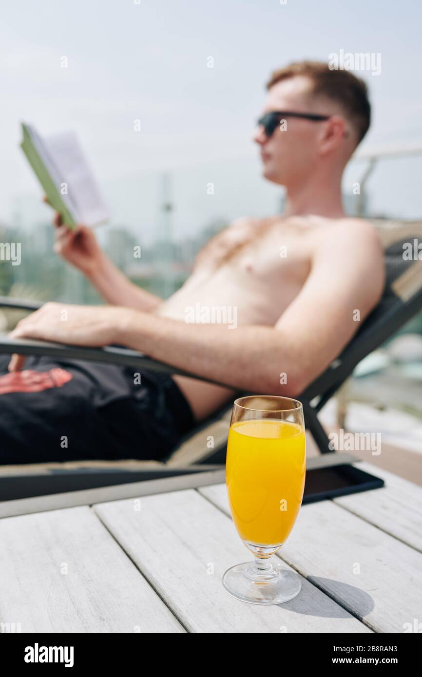 Ein Glas kalter, erfrischender Orangensaft auf dem Tisch neben dem jungen Mann, der sich in der Chaiselounge entspannen und ein Buch lesen kann Stockfoto