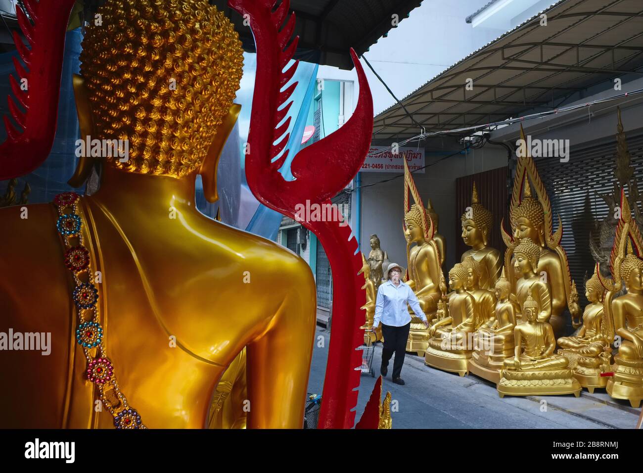 Eine Frau passiert eine Gasse in der Nähe der Bamrung Muang Road., Bangkok, Thailand, vorbei an einer Reihe von Buddha-Statuen, die in angrenzenden kleinen Fabriken hergestellt werden Stockfoto