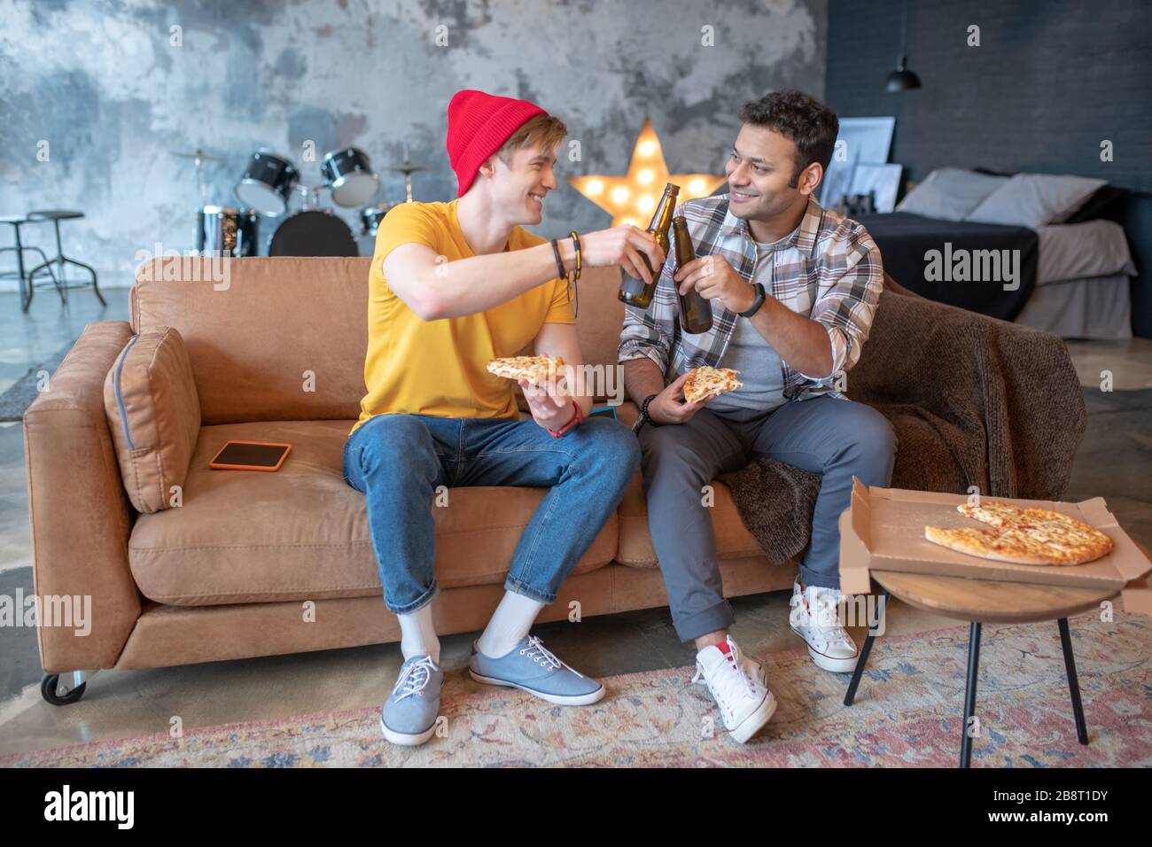 Zwei junge Männer sitzen auf dem Sofa und genießen Pizza und Bier Stockfoto