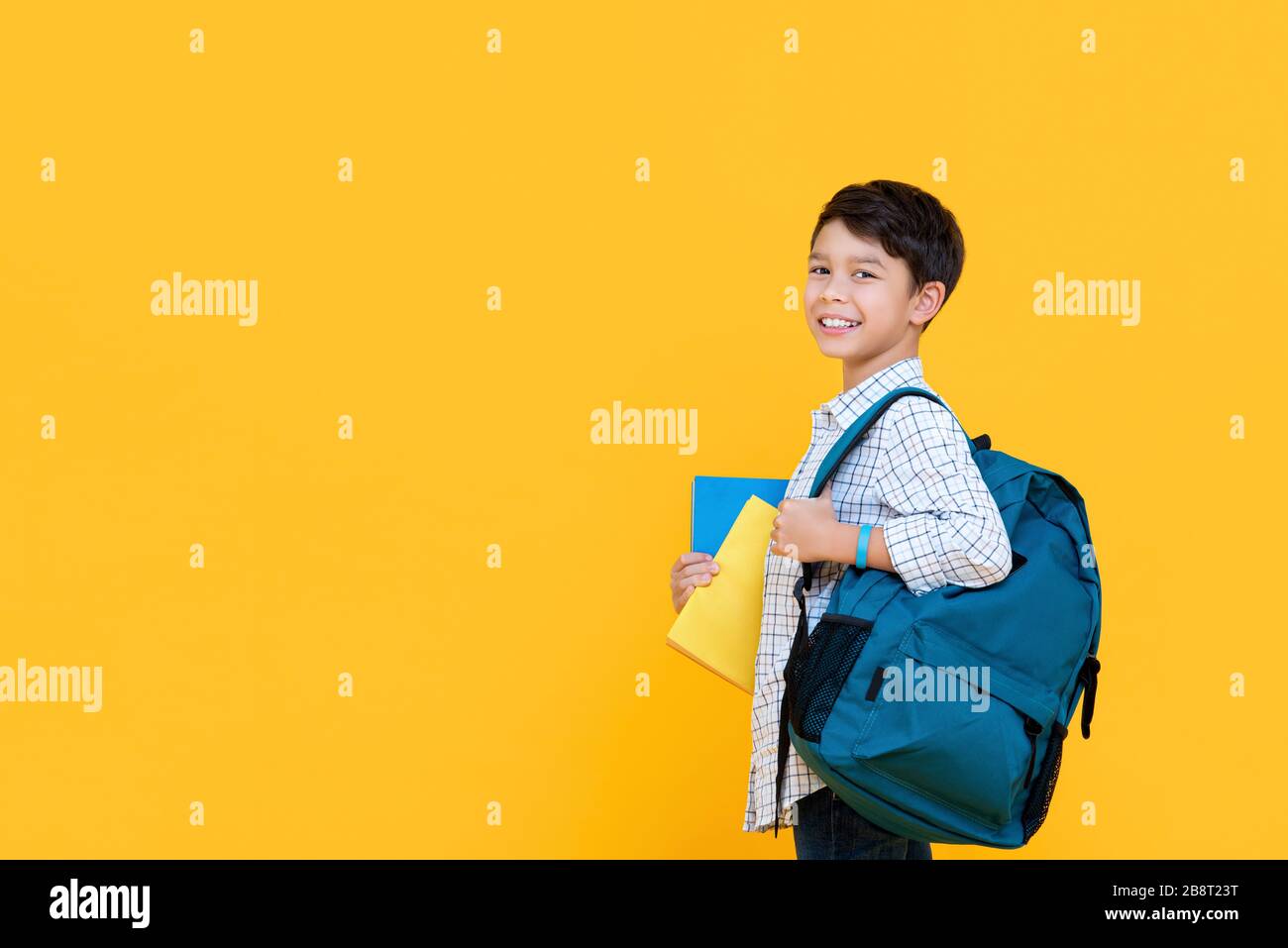 Fröhlicher lächelnder zehnjähriger gemischter Rennjunge mit Rucksack und Büchern, die bereit sind, zur Schule zu gehen, isoliert auf gelbem Hintergrund mit kopiertem Spcae Stockfoto